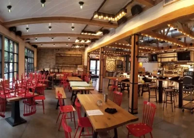 A spacious, modern dining area with wooden beams on the ceiling, red chairs upturned on tables, and a large open kitchen in the background. Empty dining tables are placed throughout the space.